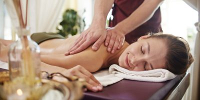 Portrait of beautiful young woman lying on massage table with eyes closed and smiling blissfully enjoying SPA treatment while man massaging her back with lotions and body oils.